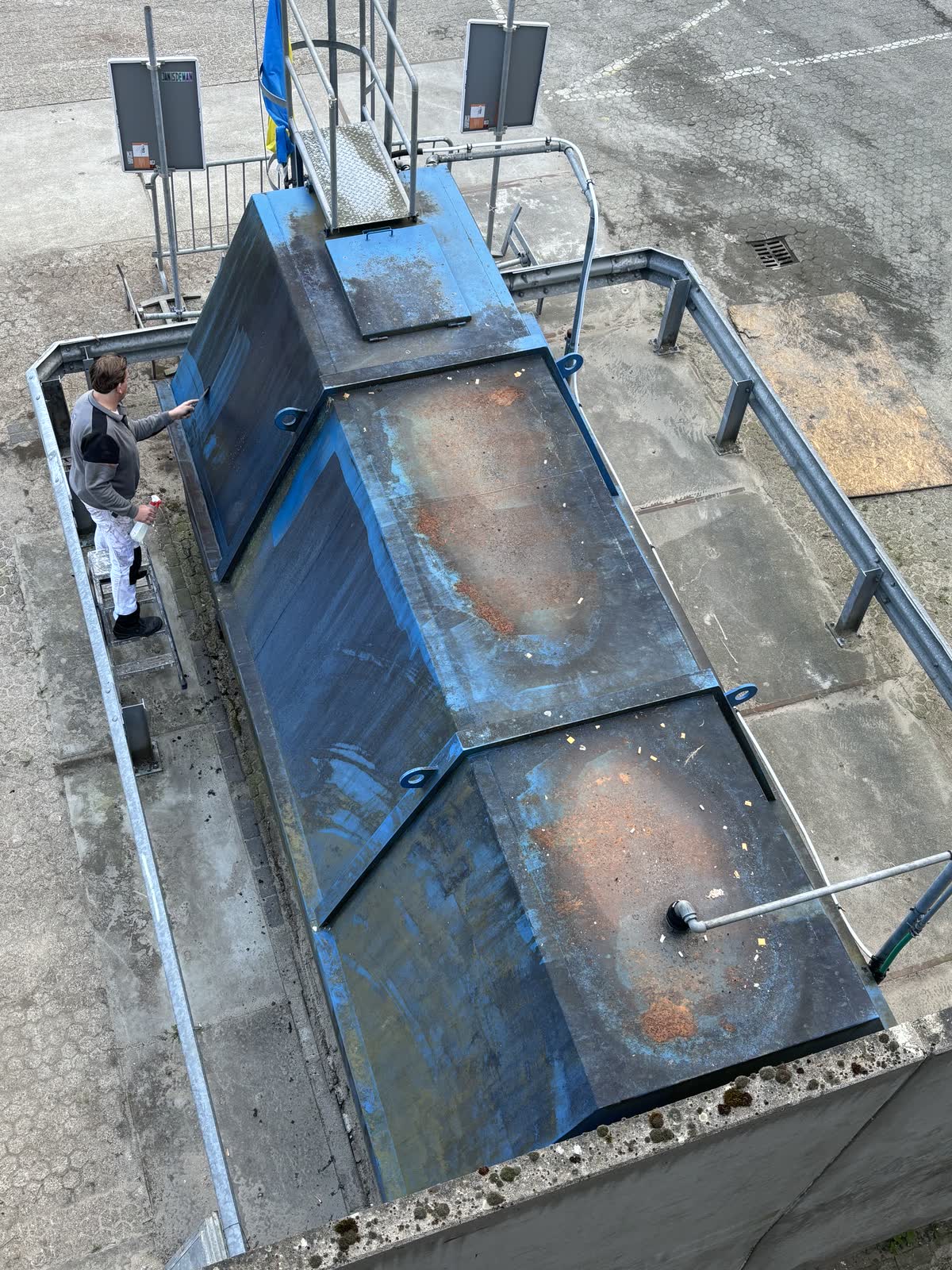 Blauwe silo van bovenaf vóór het schilderwerk, met roest en kale plekken, en een schilder ernaast